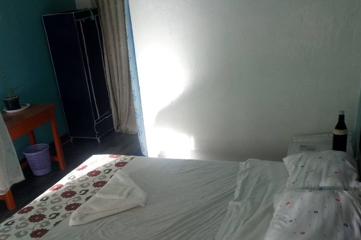 A neatly made bed with floral bedding, a small wooden table with a potted plant, and a bottle on a nightstand.