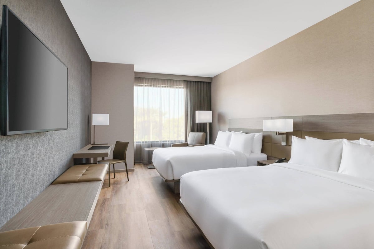 Een hotelkamer met twee bedden, een aan de muur bevestigde televisie, een bureau met stoel, en een raam met gordijnen.