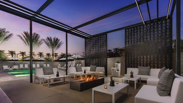 An outdoor lounge area with white sofas, a fire pit, and a pool in the background.