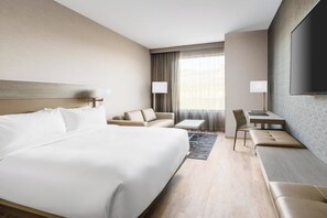 Habitación de hotel con una cama grande, un sofá, una mesita, una silla y un televisor fijado a la pared.
