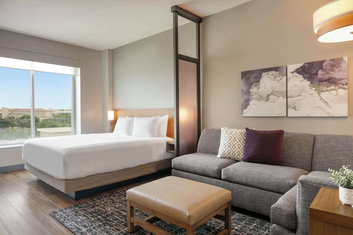 A hotel room with a large bed, a grey sofa, a wooden bench, and a view of the city through large windows.