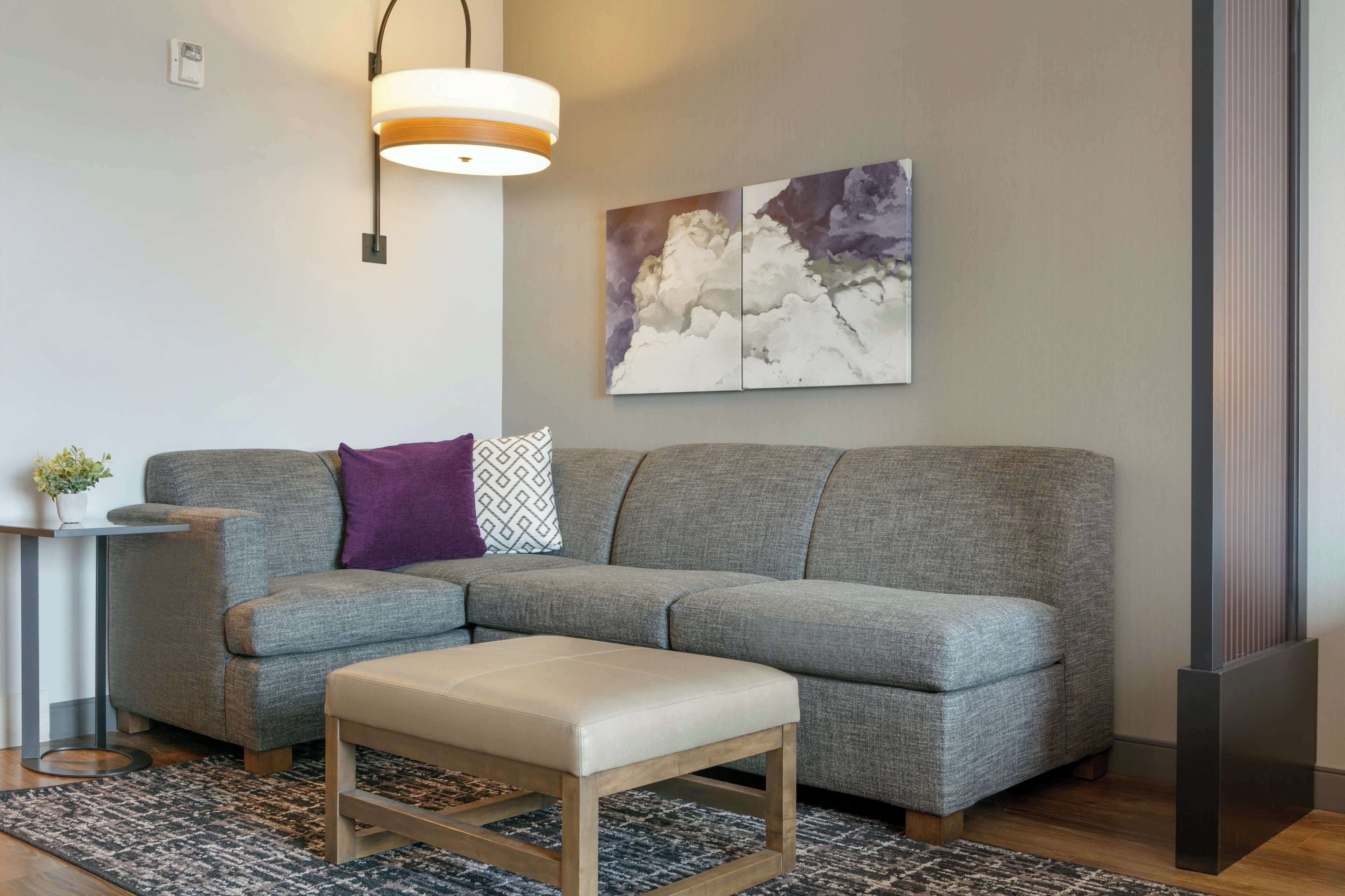 Una sala moderna con un sofá seccional gris, una mesita auxiliar con una planta, un banco de madera, un tapiz estampado y un cuadro abstracto colgado en la pared.