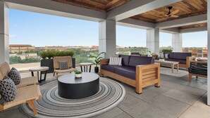 Un vaste espace de détente extérieur doté d’un plafond en bois, de mobilier moderne et de grandes fenêtres offrant une vue sur la ville.