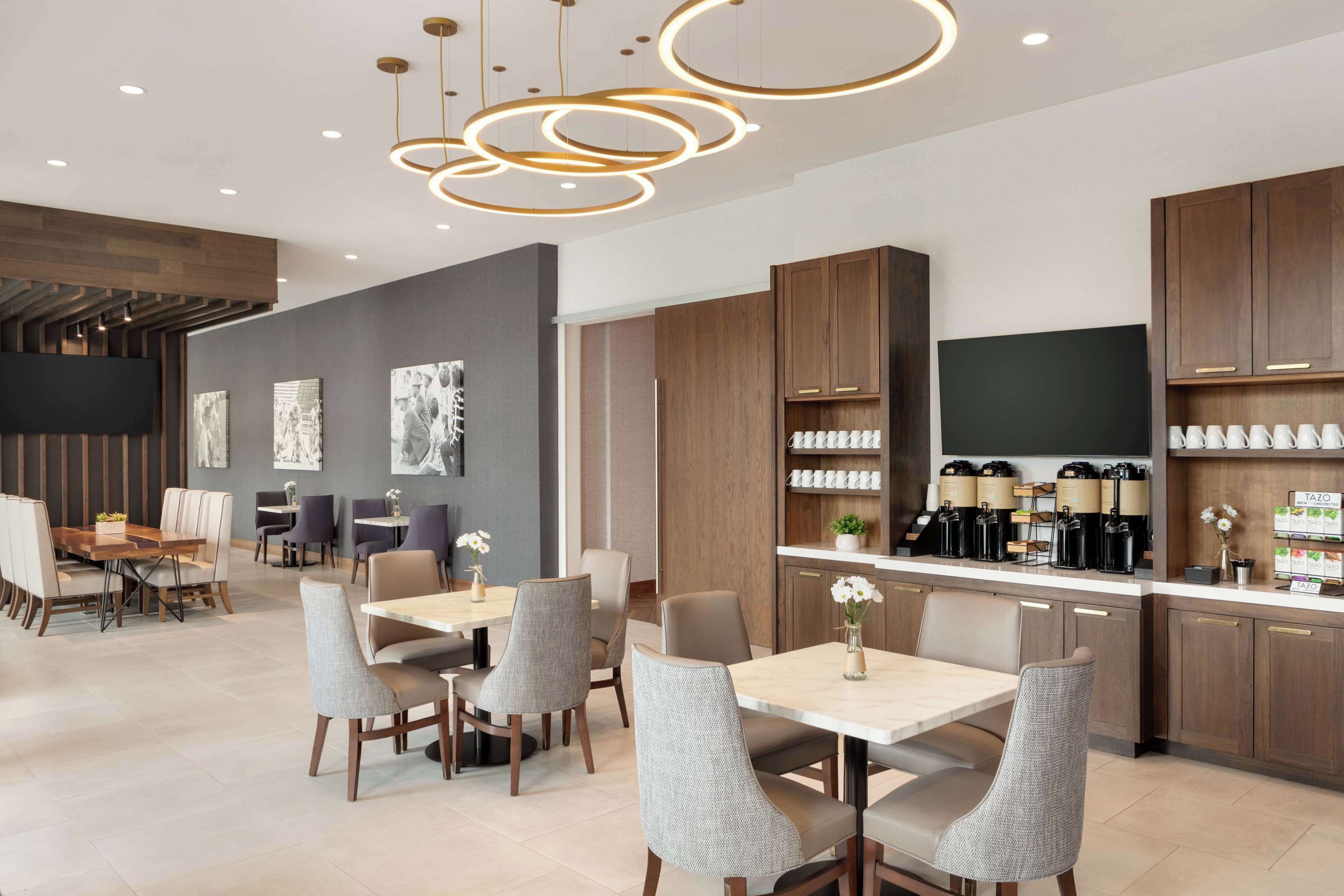 A modern hotel dining area with a long communal table, individual dining tables, and a bar area with coffee machines.