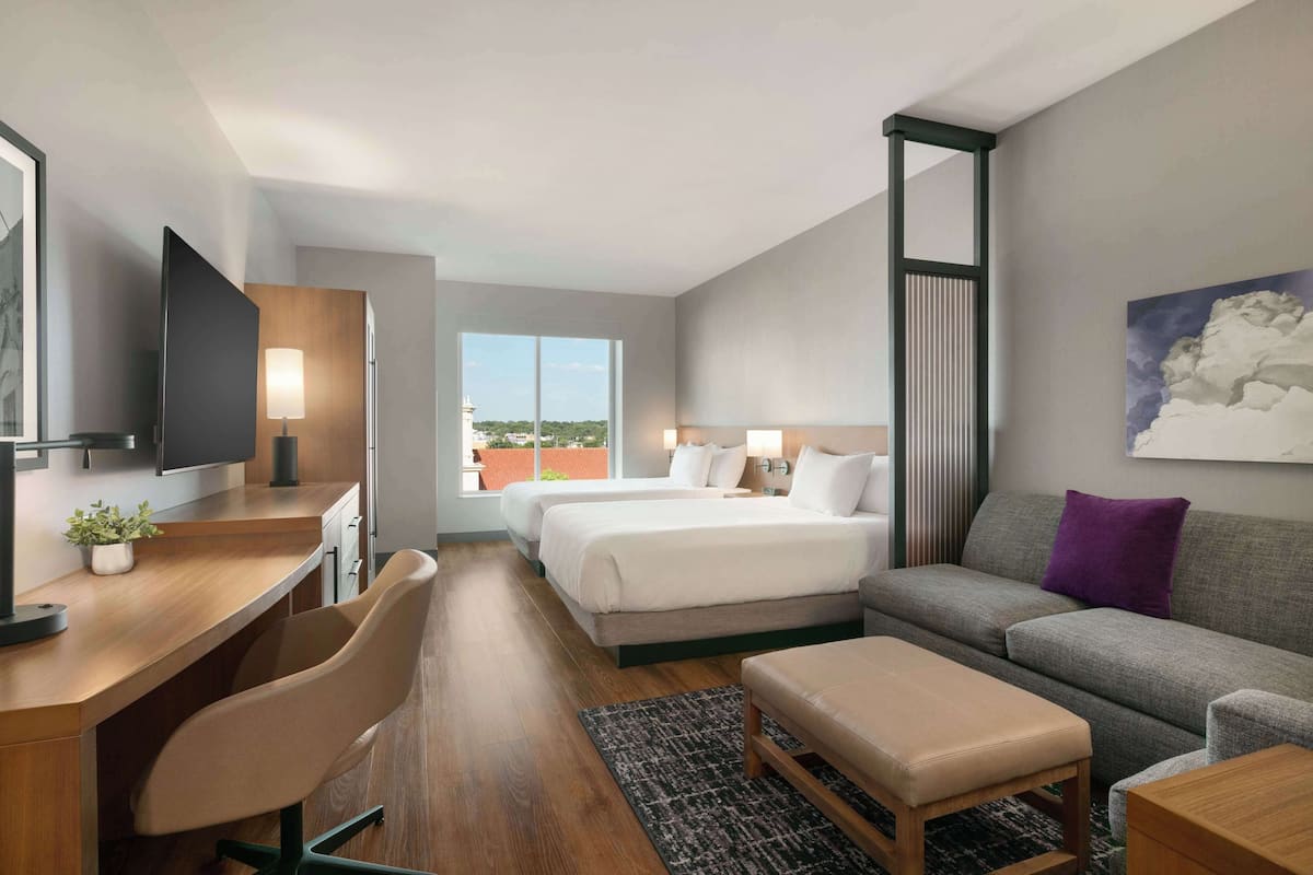 A hotel room with a desk, two beds, a sofa, a TV, and a large window with a view.