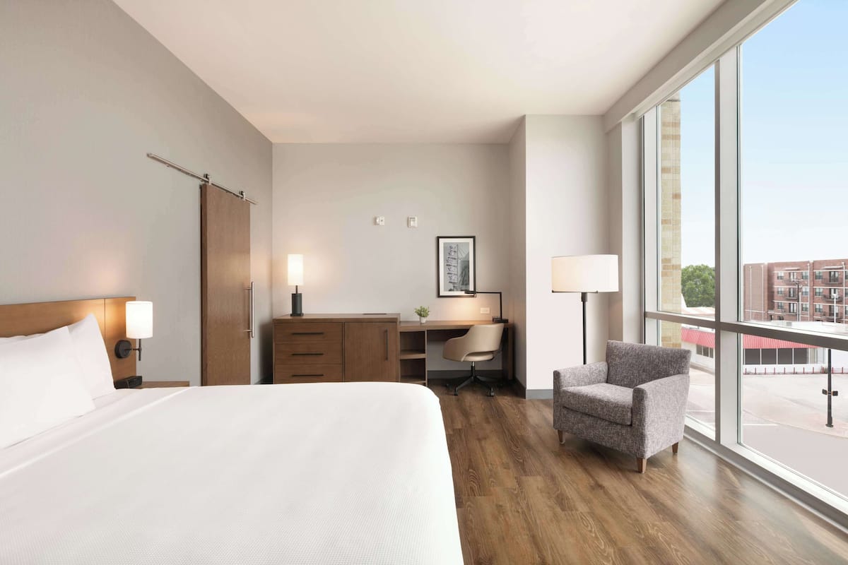 A hotel room with a large bed, a desk, a chair, and a view through a large window.