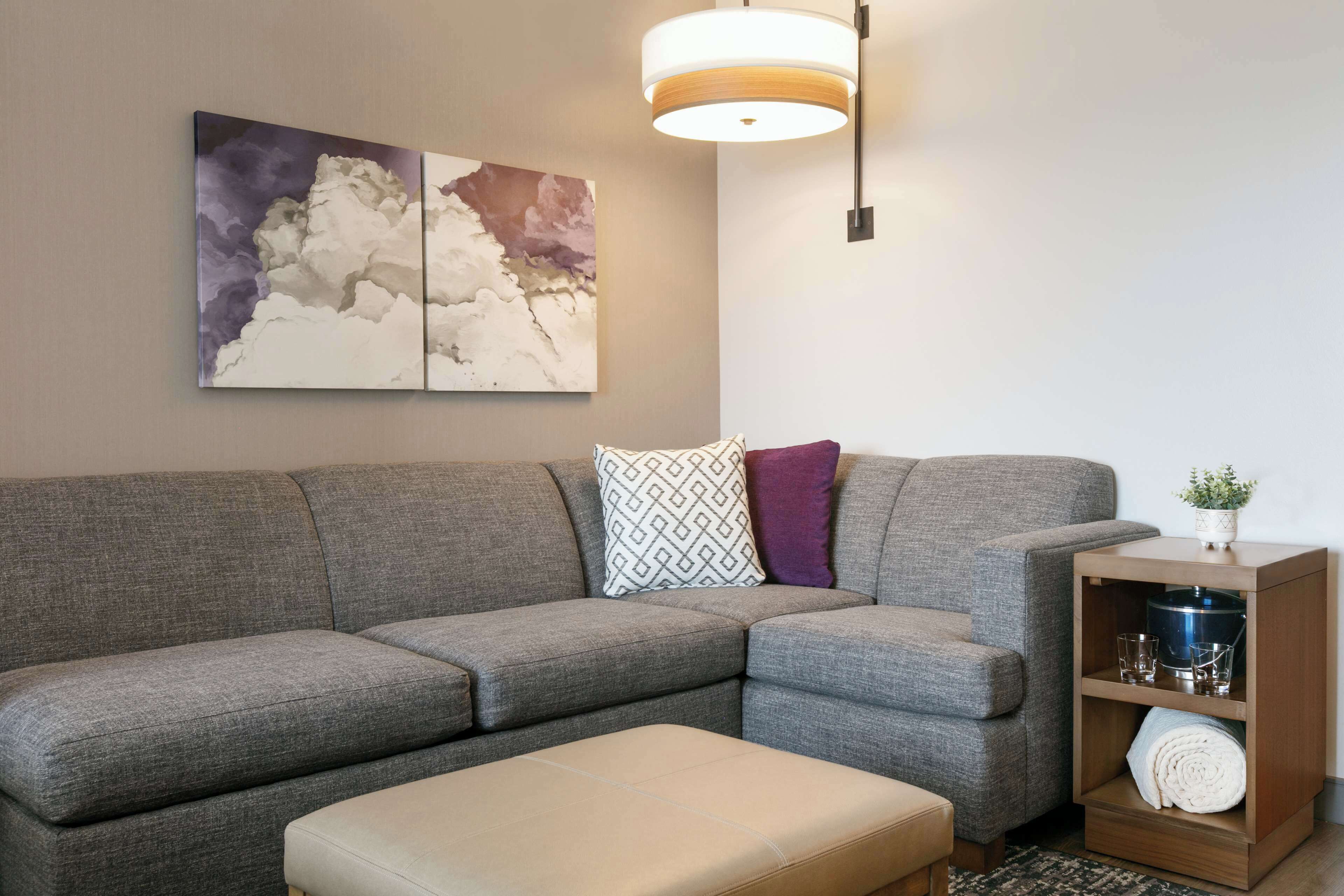 Uma sala de estar moderna com um sofá seccional cinza, um banquinho combinando, uma mesa lateral de madeira com uma planta e uma pintura abstrata pendurada na parede.