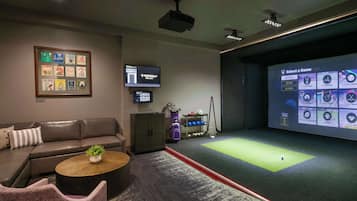 A room with a green carpeted area, a brown leather sofa, a wooden coffee table, a framed picture on the wall, a television, and a projector screen displaying game options.