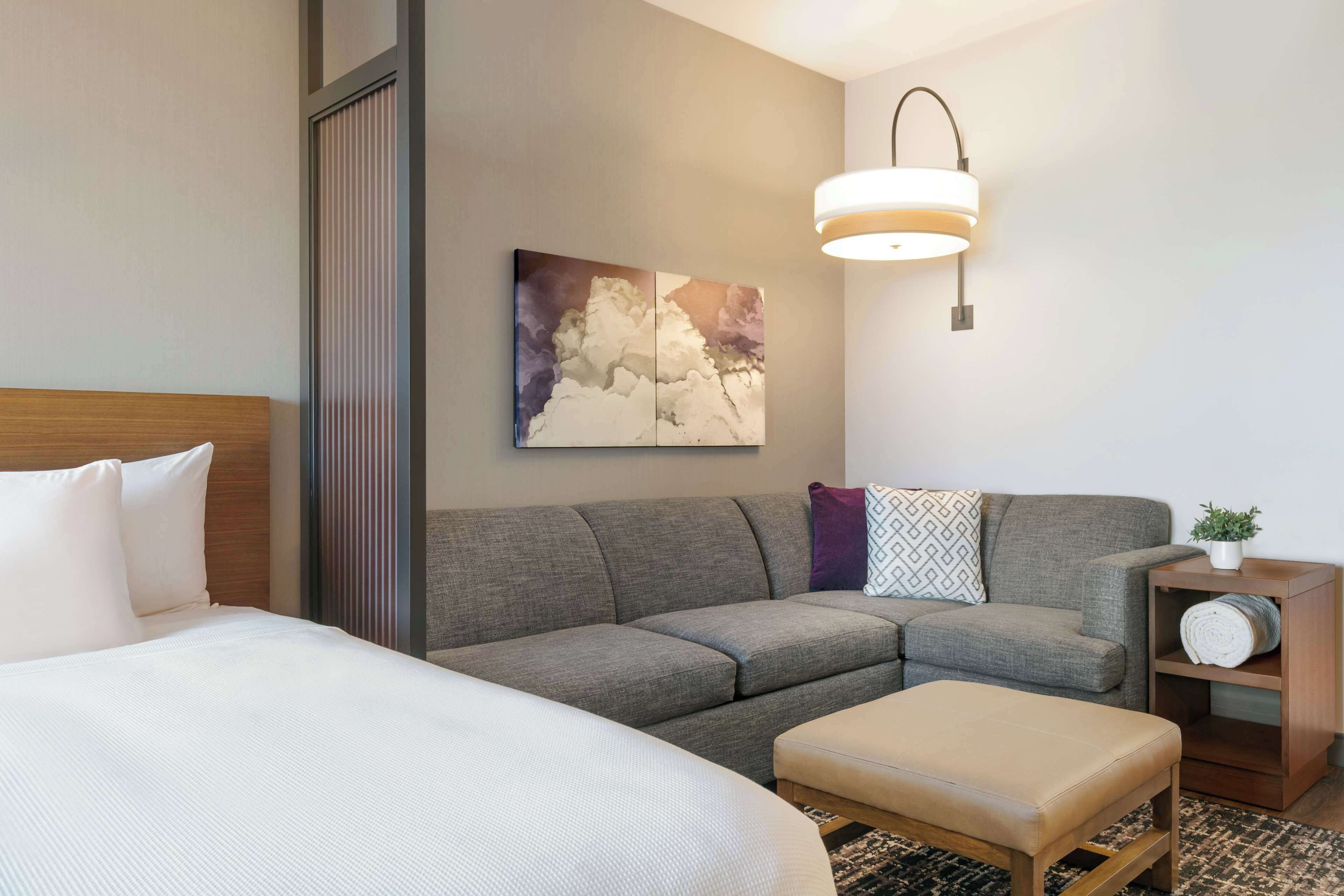 A hotel room with a bed, a gray sofa, a small wooden side table with a plant, and a wall-mounted light fixture.
