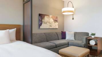 A hotel room with a bed, a gray sofa, a small wooden side table with a plant, and a wall-mounted light fixture.