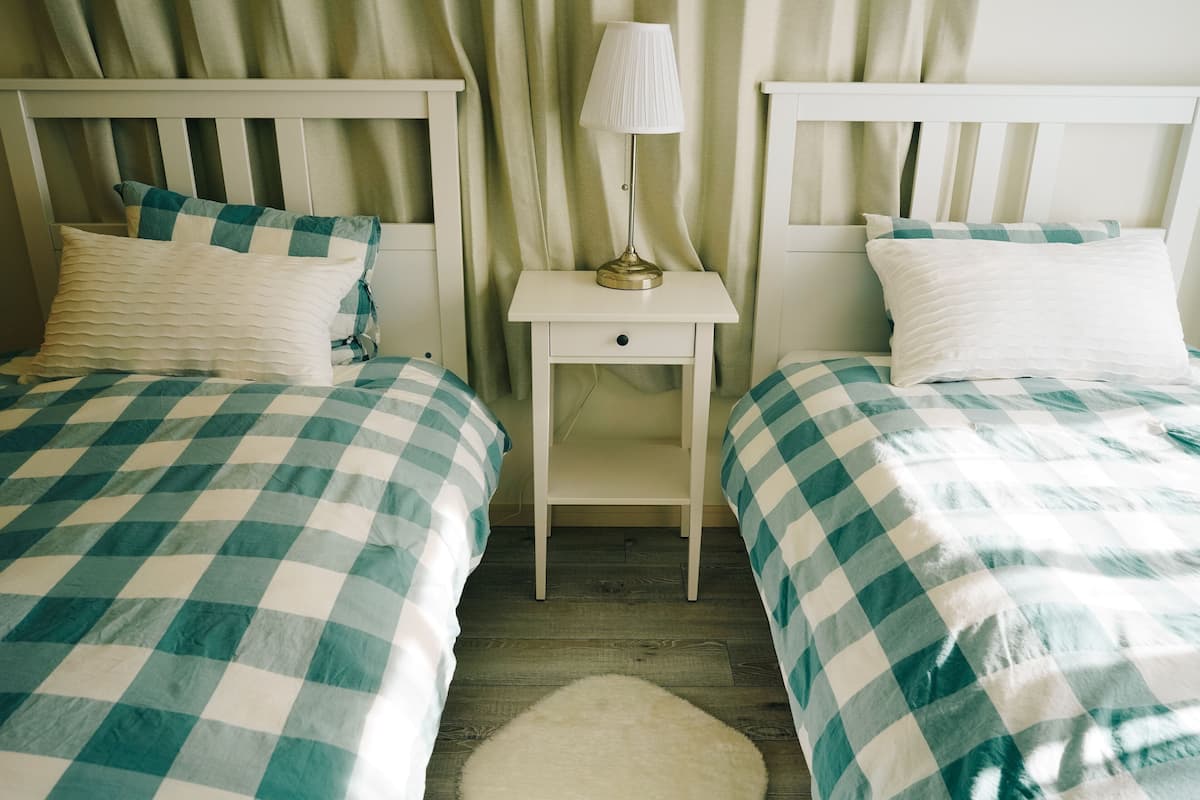 Two twin beds with checkered bedding, a bedside table with a lamp, and a rug on a wooden floor.