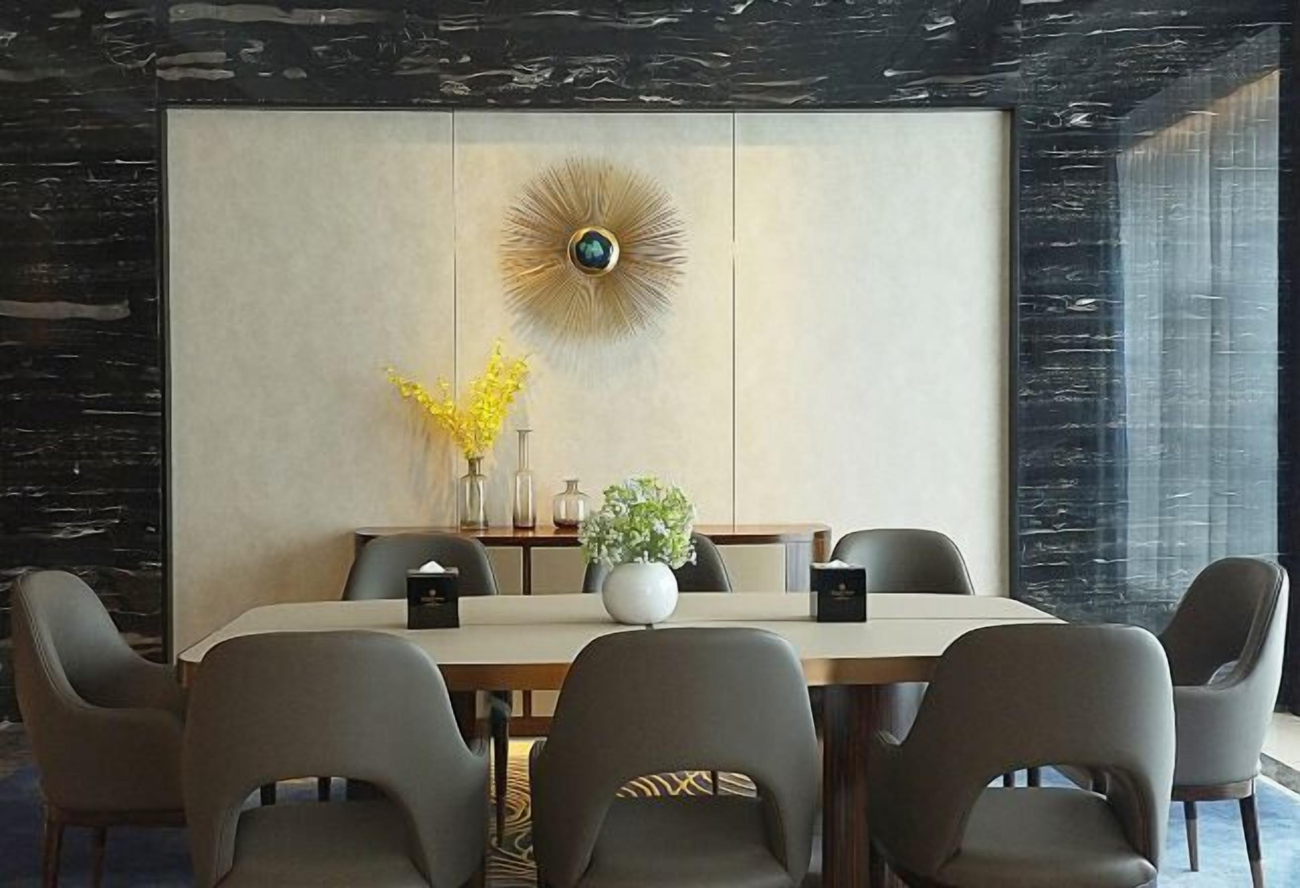 A modern dining area with a large table, six chairs, a decorative wall piece, and a vase with yellow flowers.
