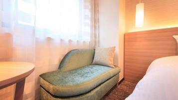 A hotel room with a bed, a small sofa, a bedside table, and a window with sheer curtains.