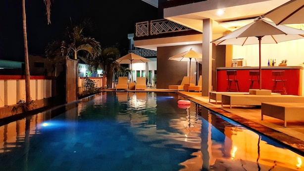 A nighttime swimming pool area with lounge chairs and umbrellas, a bar area with stools, and a staircase leading to an upper level. - Talent Resort (Gili Trawangan)