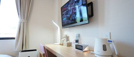 A hotel room with a wooden desk, a flat-screen TV mounted on the wall, a desk lamp, a digital clock, a kettle, and a chair.
