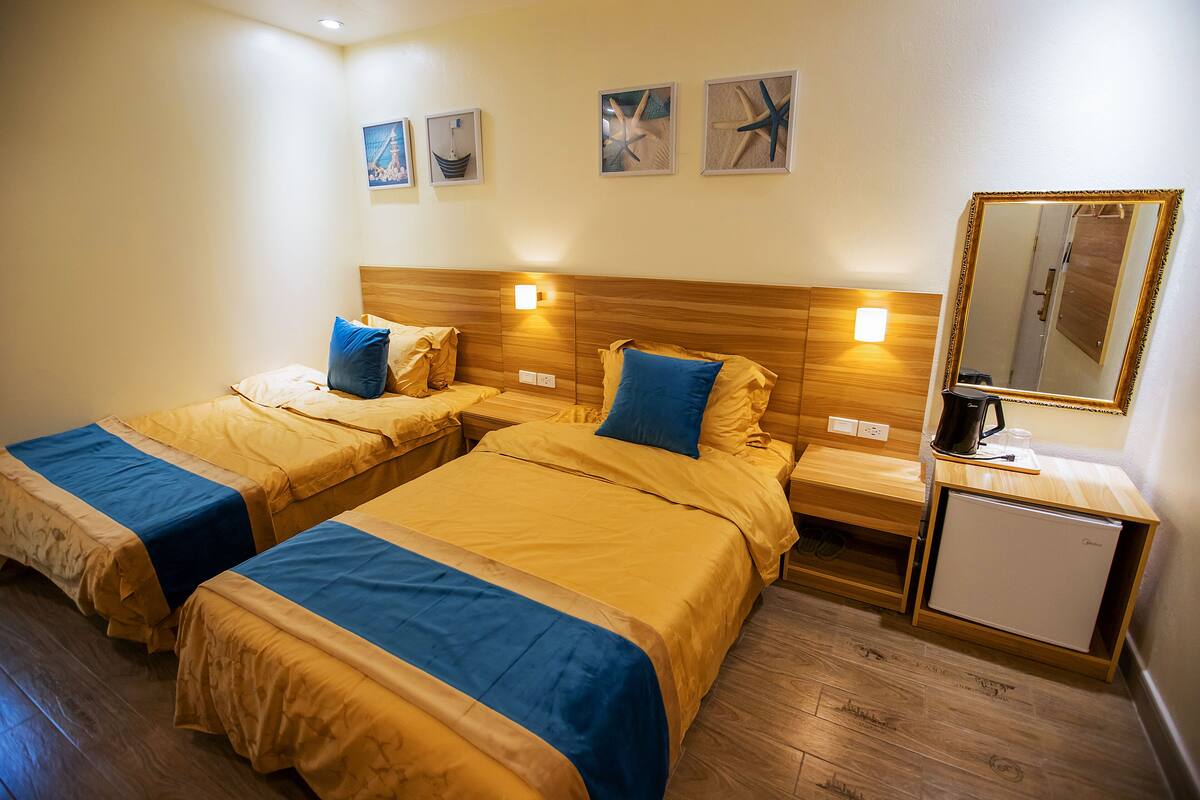 Habitación de hotel con dos camas, cabeceros de madera, mesitas de noche, un refrigerador pequeño y una obra de arte colgada en la pared.