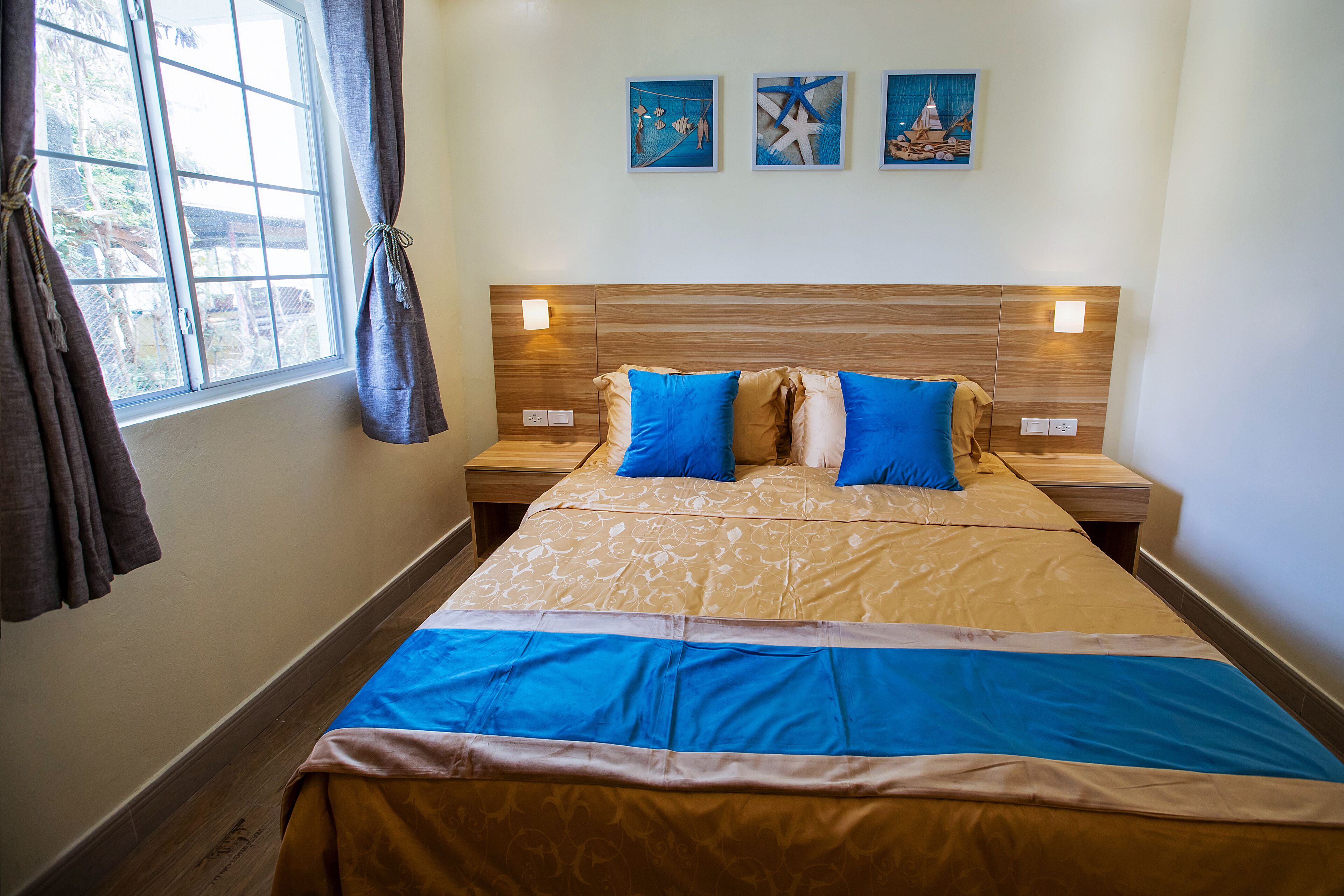 A hotel room with a large bed, two bedside tables, a window with curtains, and three wall-mounted paintings.