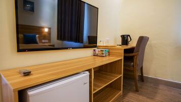 A hotel room with a large flat-screen TV mounted on the wall, a wooden desk with a chair, a small white refrigerator, and a coffee maker on the desk.