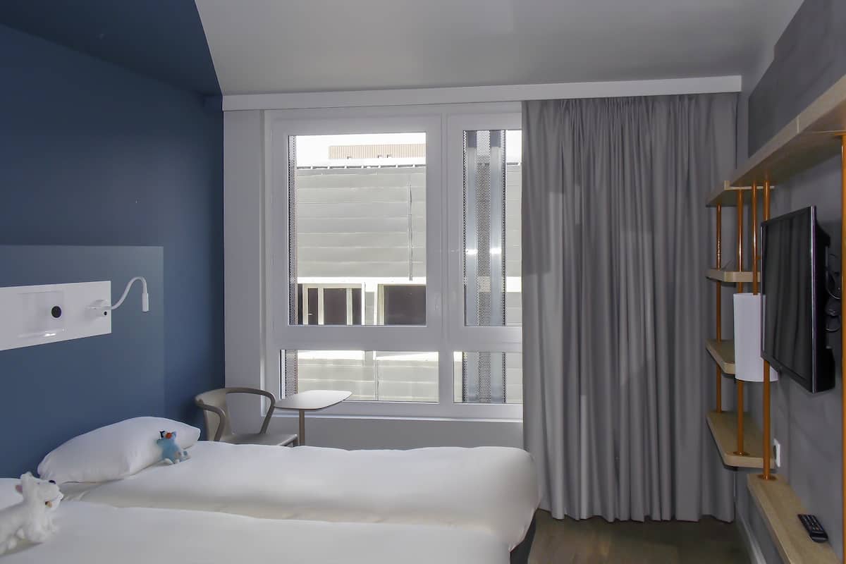 A hotel room with a bed, a small table, a chair, a TV on a wooden shelf, and a window with curtains.