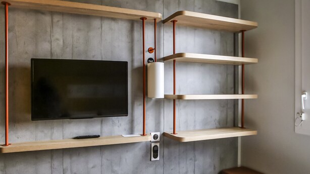 A modern room with a mounted TV, wooden shelves, and a small table. - ibis budget Rouen Centre Rive Gauche (Rouen)