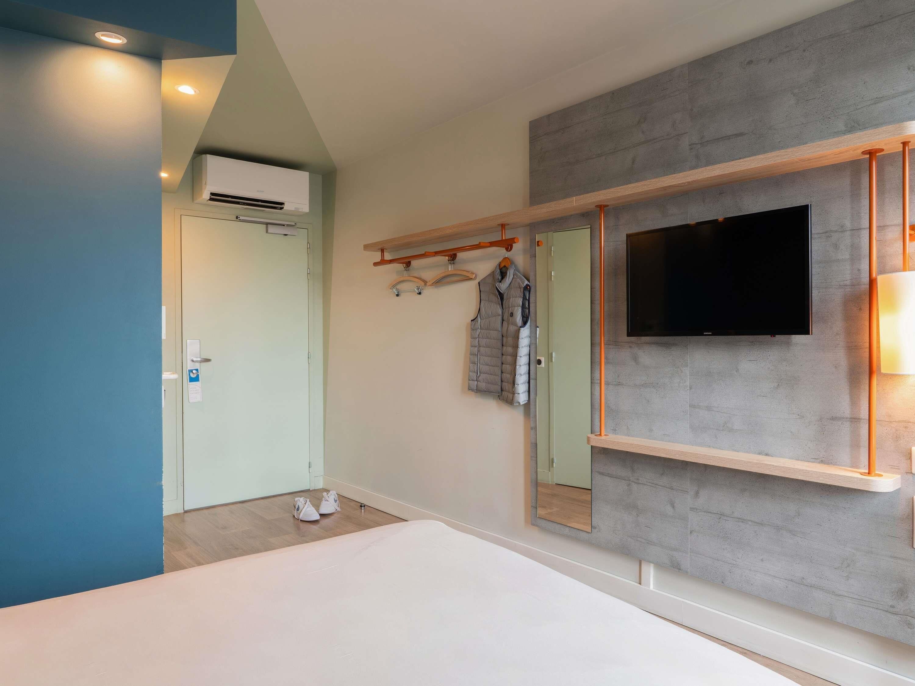 Habitación de hotel con cama, televisión montada en una pared gris, perchero con chaquetas y equipo de aire acondicionado.