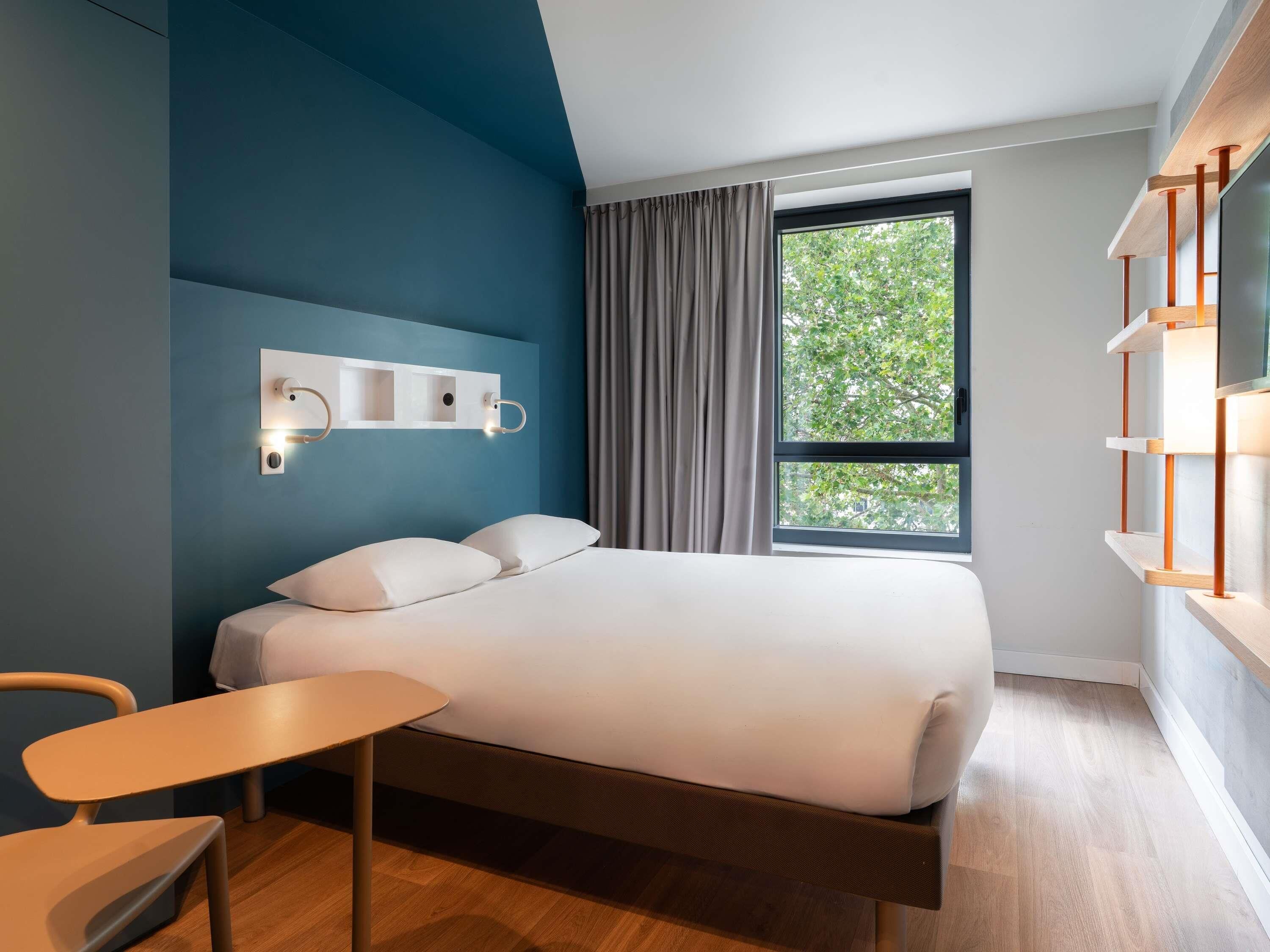 A modern hotel room with a bed, a small table, and a chair. There is a window with curtains and a view of trees outside.