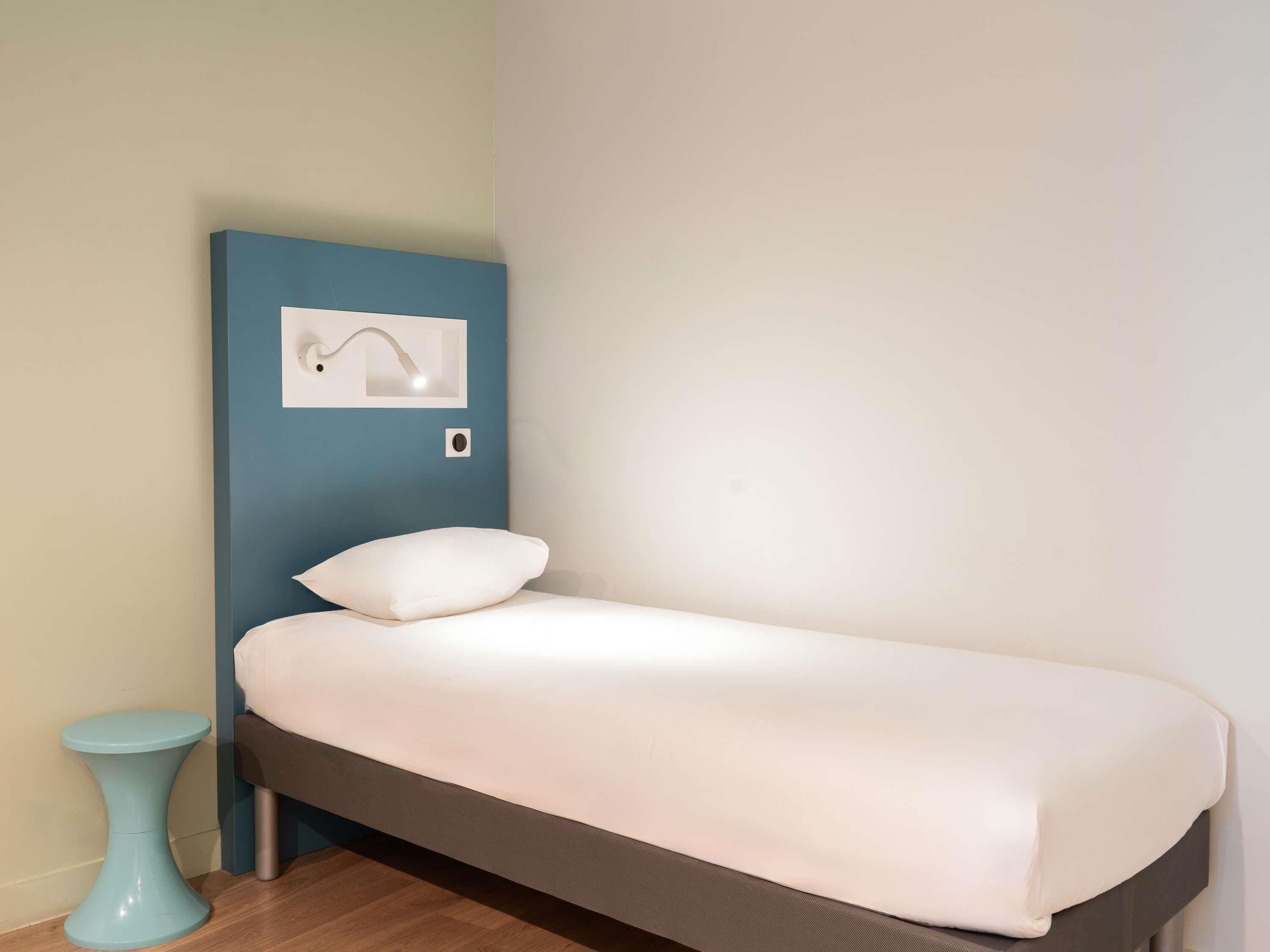 Una cama individual con colchón y almohada blancos, una mesita de noche azul y una lámpara de pared.