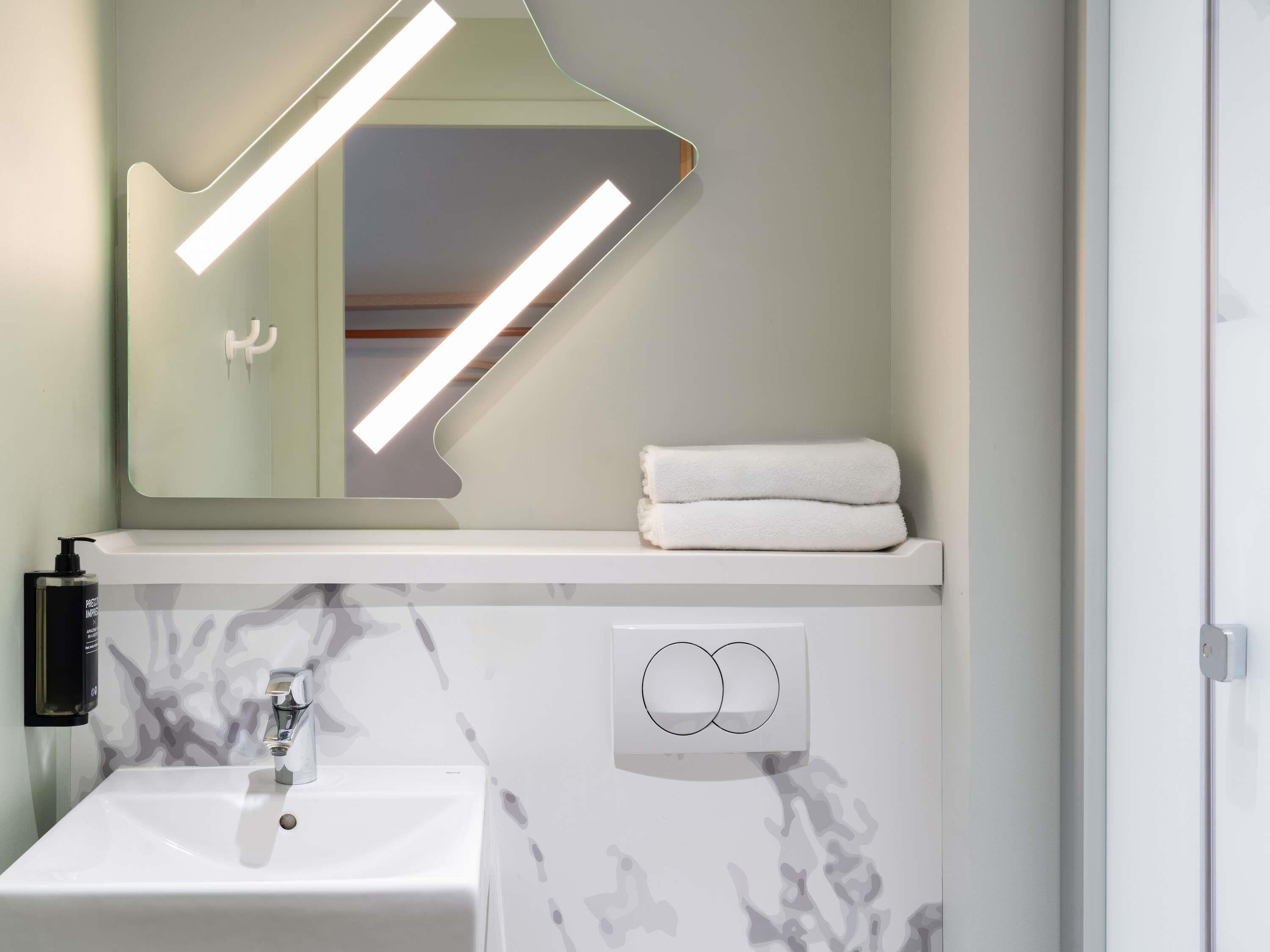 A bathroom with a white sink, a mirror with integrated lighting, a towel rack, and a soap dispenser.