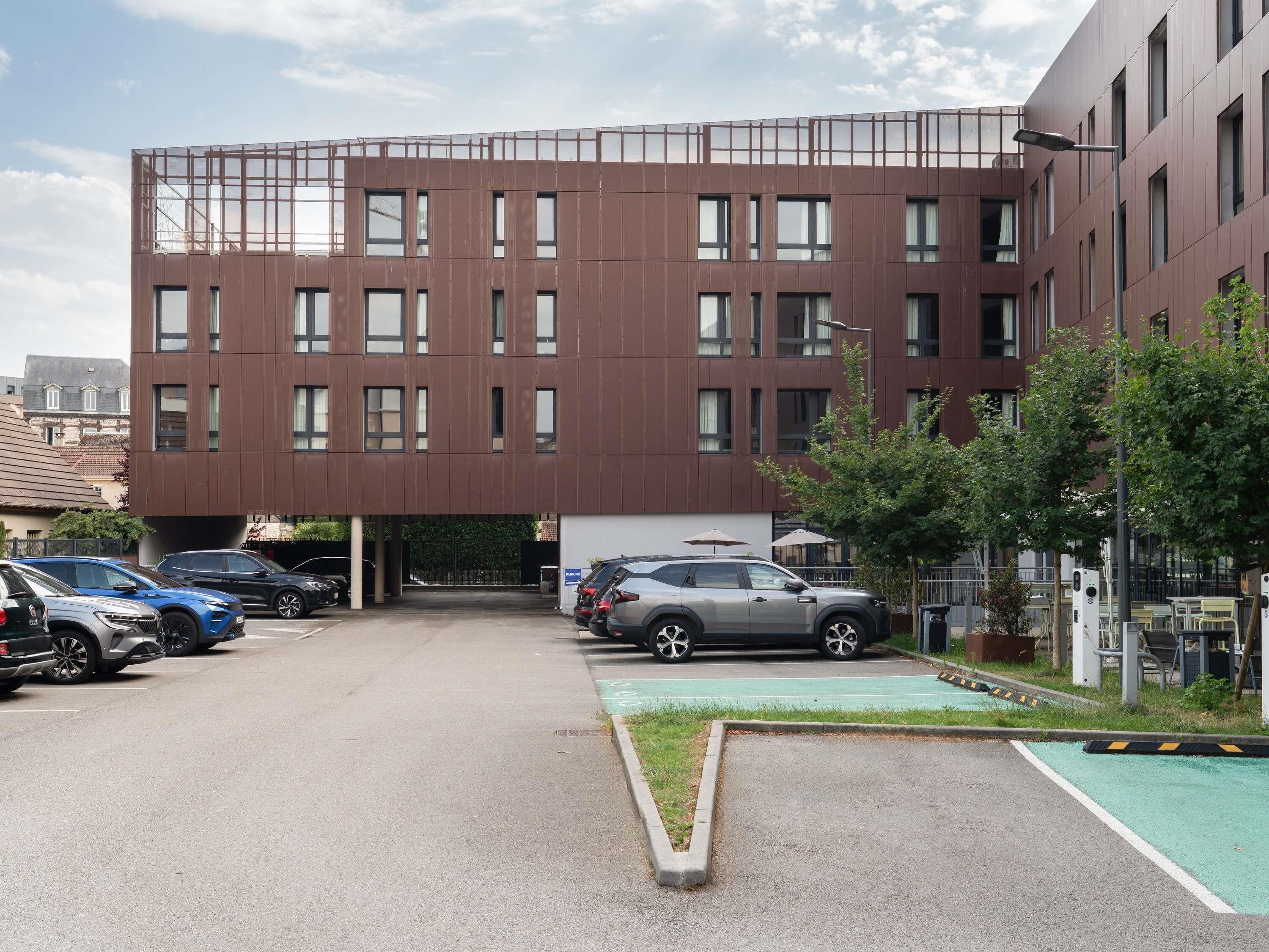 Un bâtiment moderne à plusieurs étages, un espace de stationnement avec plusieurs voitures et un petit espace vert avec des arbres.