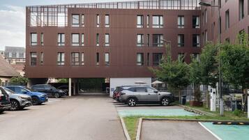 A modern building with multiple floors, a parking area with several cars, and a small green space with trees.