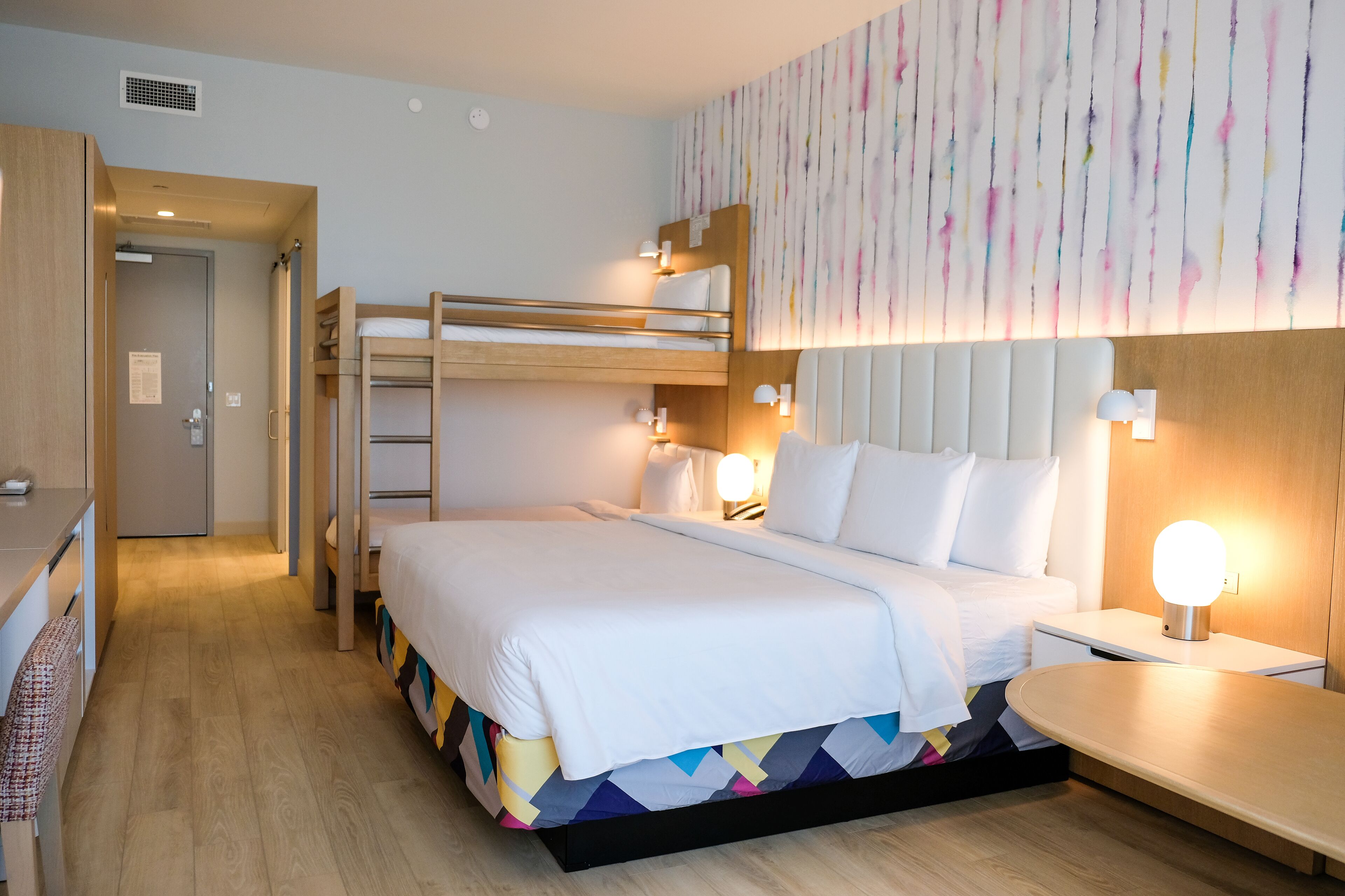 A hotel room with a bunk bed, a desk, and a bedside table with a lamp.