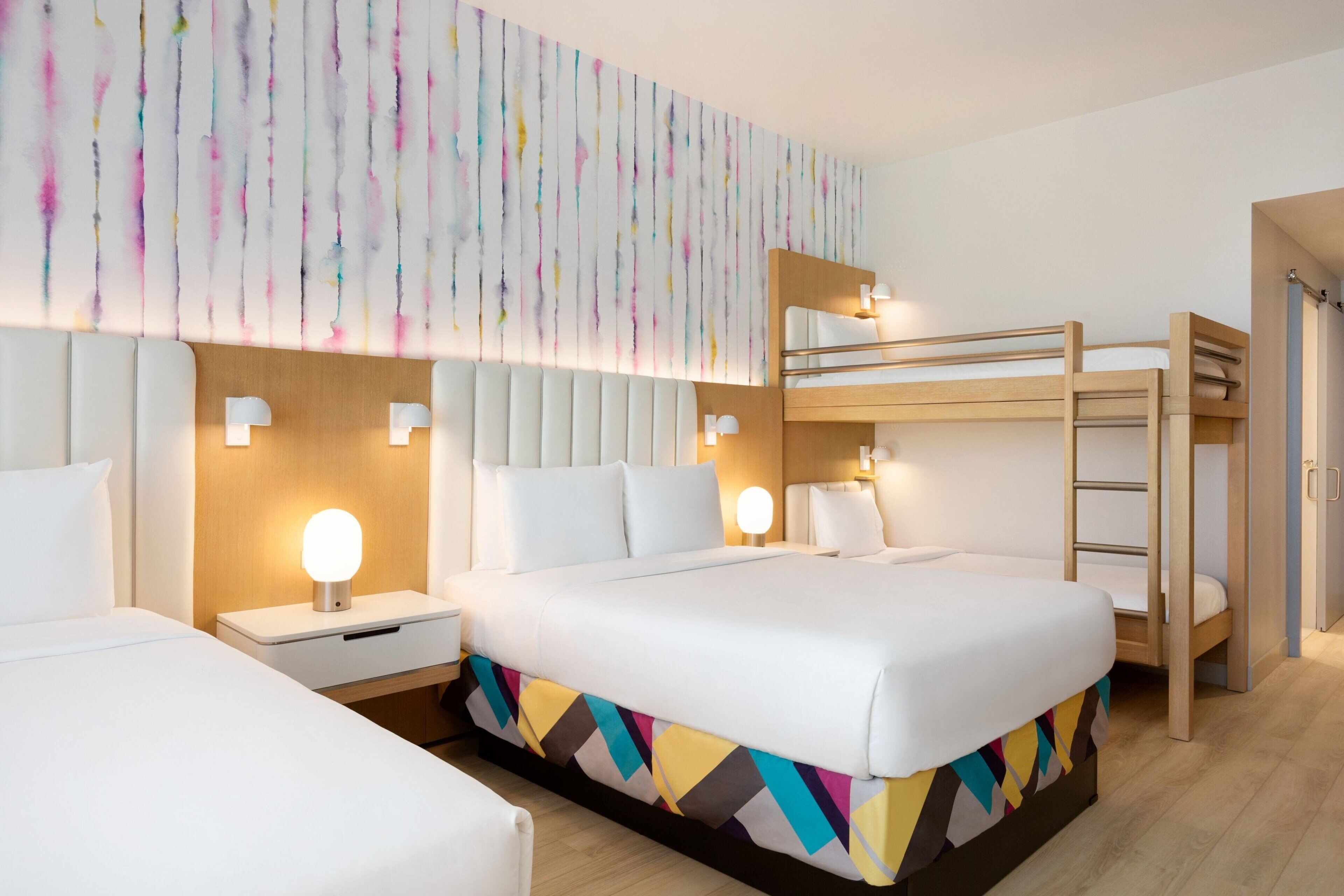 Ein modernes Hotelzimmer mit einem großen Bett, einem Etagenbett und einem farbenfrohen Bettlaken mit geometrischem Muster.
