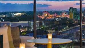 Vue depuis une chambre d’hôtel, avec une grande fenêtre donnant sur un paysage urbain au crépuscule, où l’on aperçoit des attractions illuminées d’un parc d’attractions et des bâtiments.