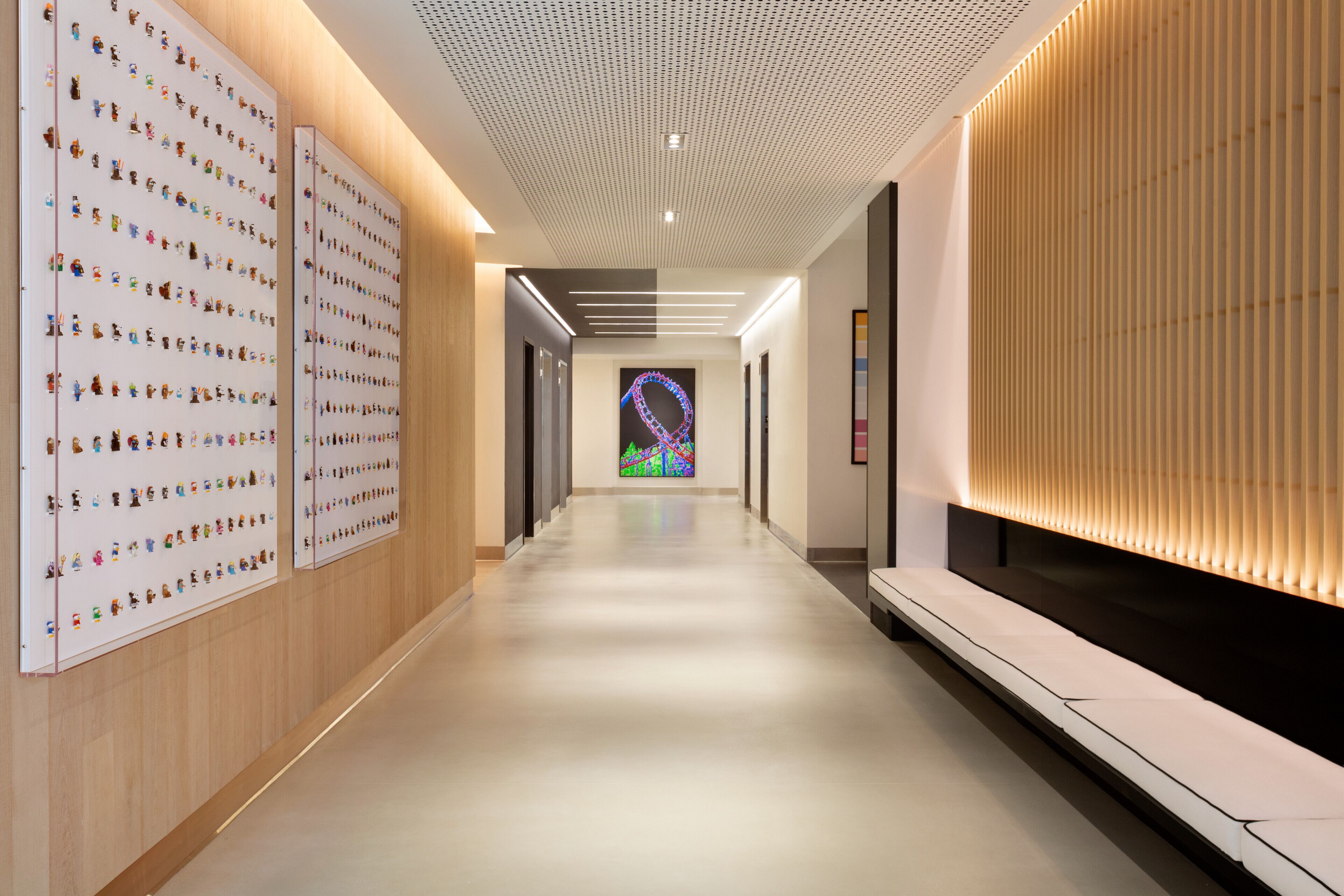 A modern hallway with a long, straight layout, featuring a wall with numerous small figures and a large, colorful artwork at the end.