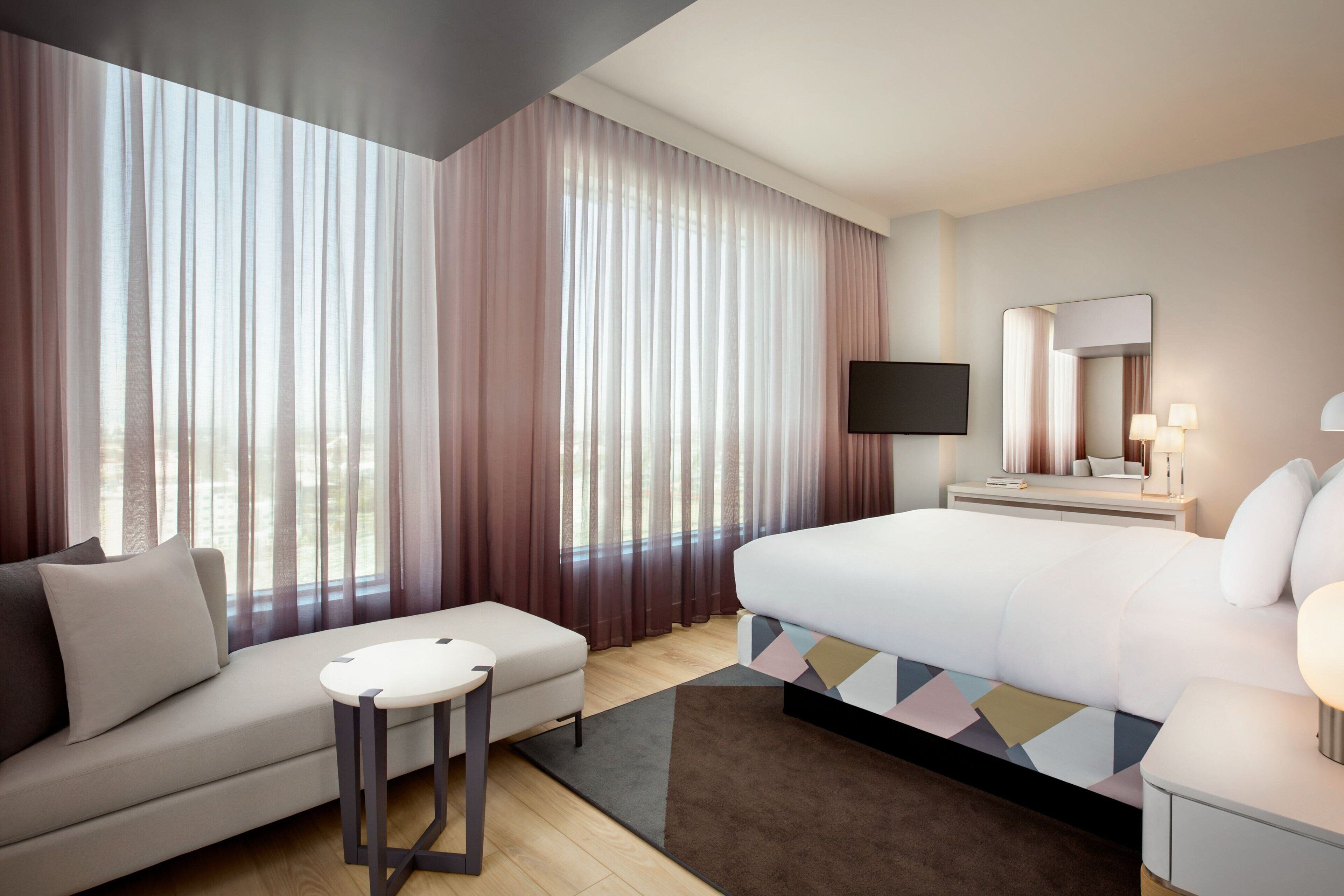A hotel room with a bed, a sofa, a small table, a TV, and a large window with curtains.