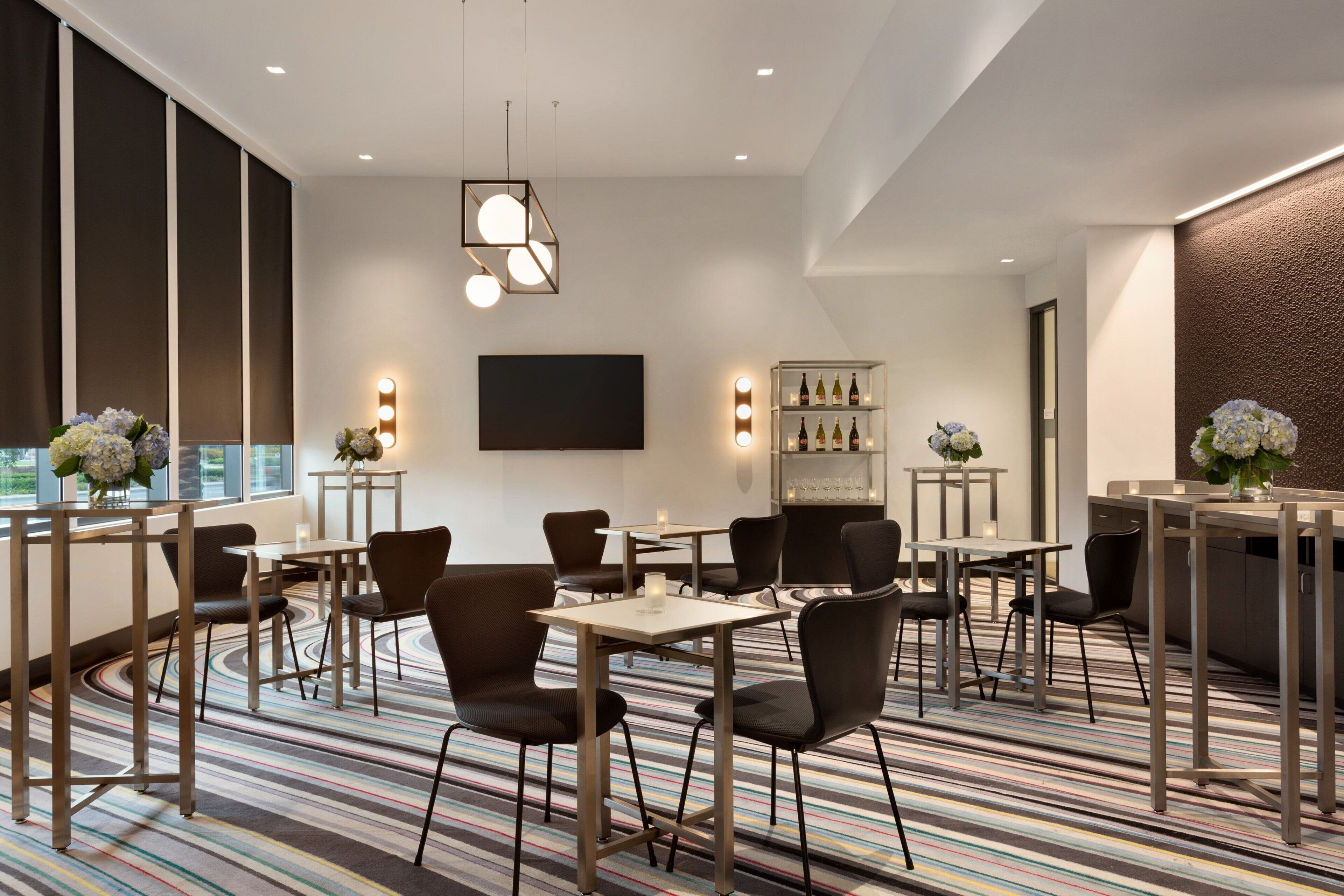 A modern hotel dining area with striped carpet, multiple tables and chairs, a bar area, and a large TV screen.