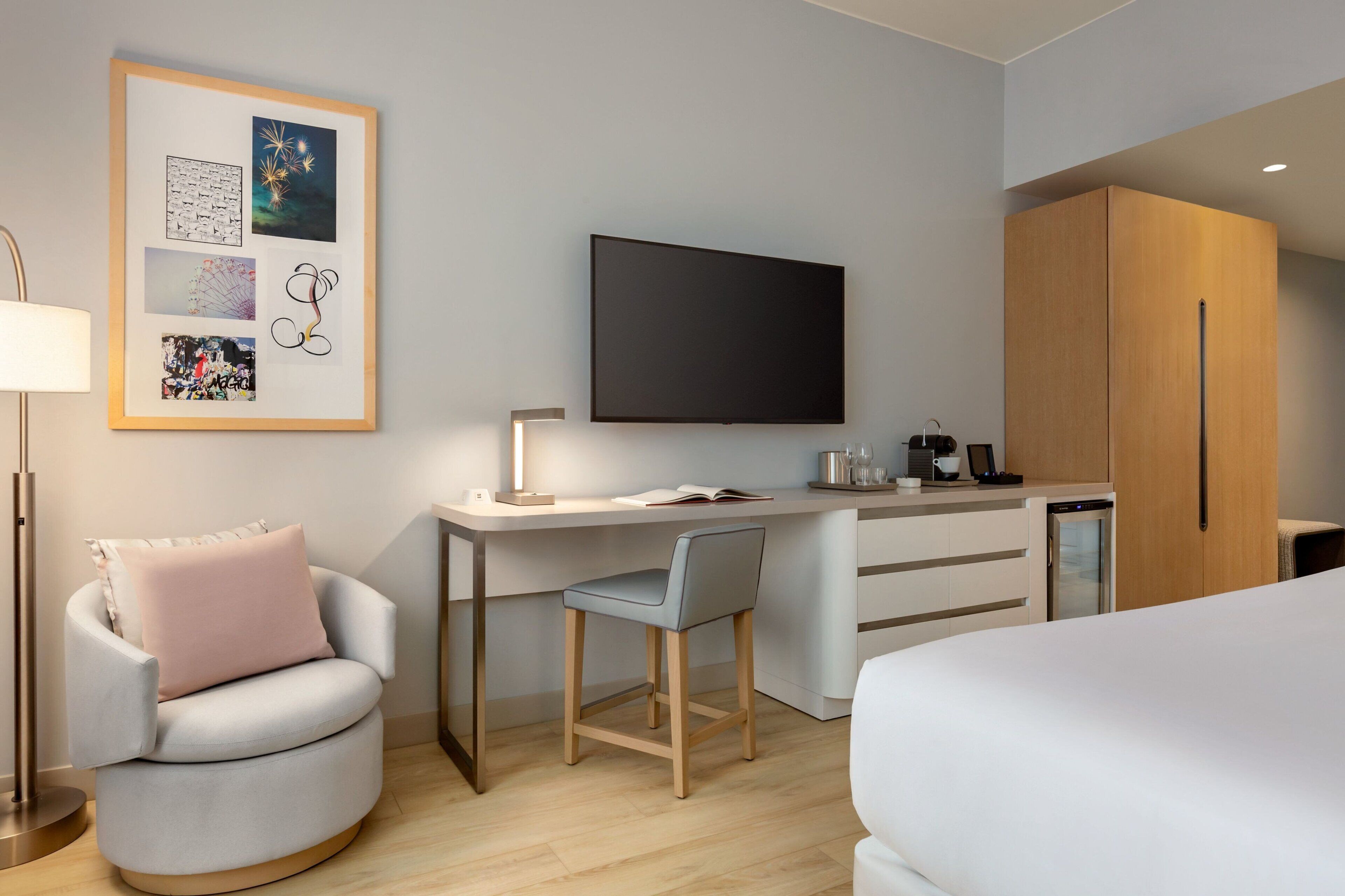 A modern hotel room with a curved sofa, a desk with a flat-screen TV, a kitchenette, and a bed.