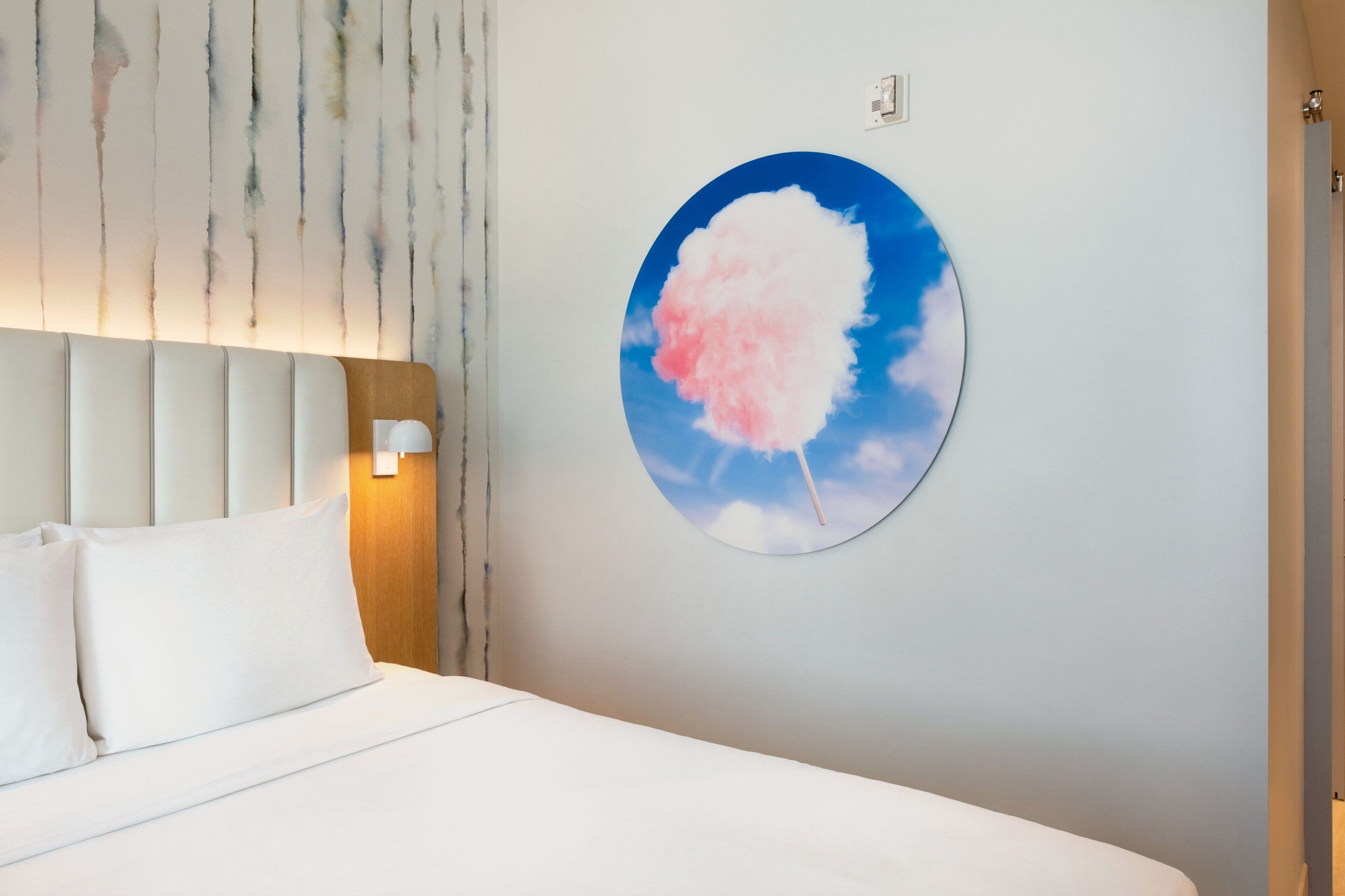 Una habitaci ón de hotel con cama, cabecera, un cuadro colgado en la pared de una nube de algodón de azúcar y un interruptor de luz.