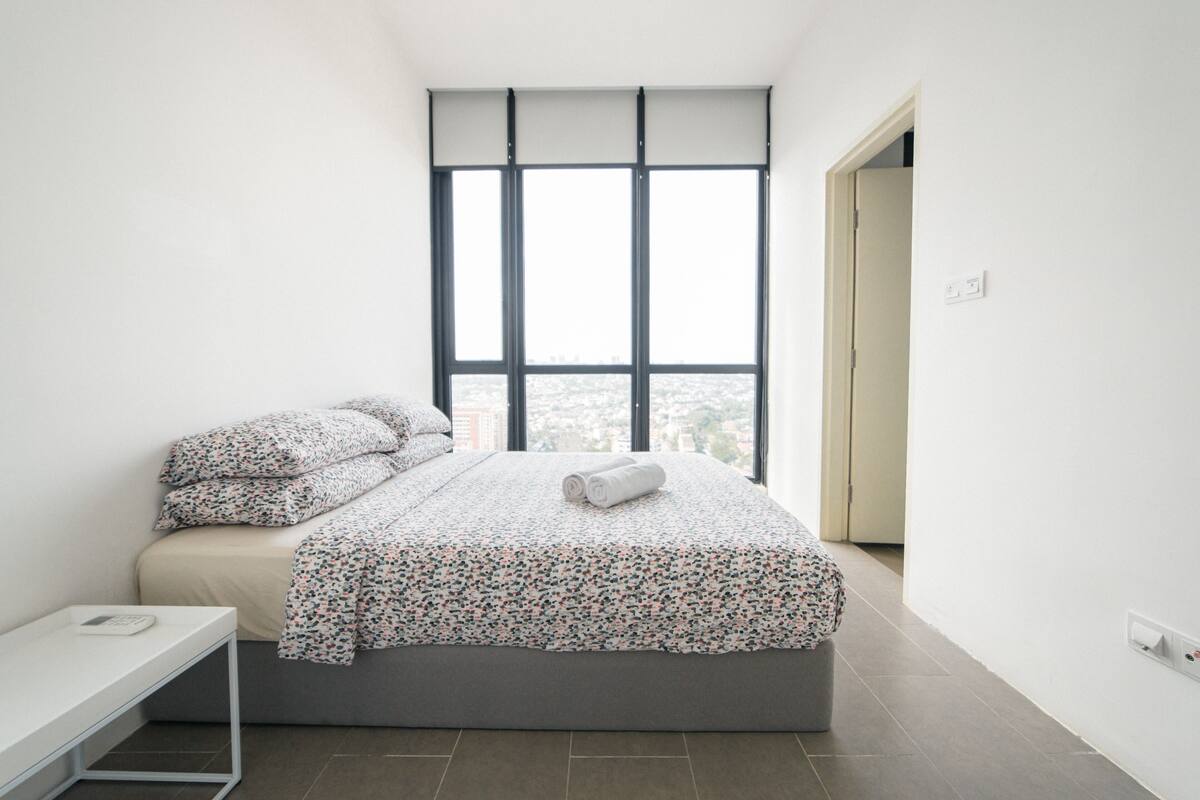 A modern bedroom with a large window, a bed with floral bedding, a bedside table, and a view of the city.