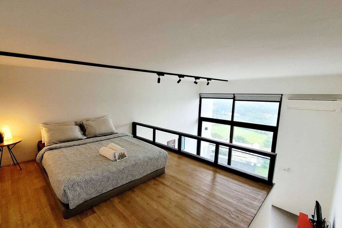 A bedroom with a large bed, a small table with a lamp, and a window with a view of buildings.
