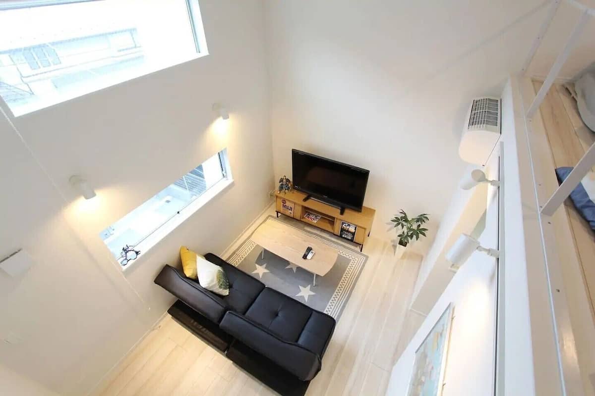 Una sala moderna con un sofá negro, un televisor de pantalla plana y una mesita. Hay una ventana que proporciona luz natural y una planta en el alféizar.