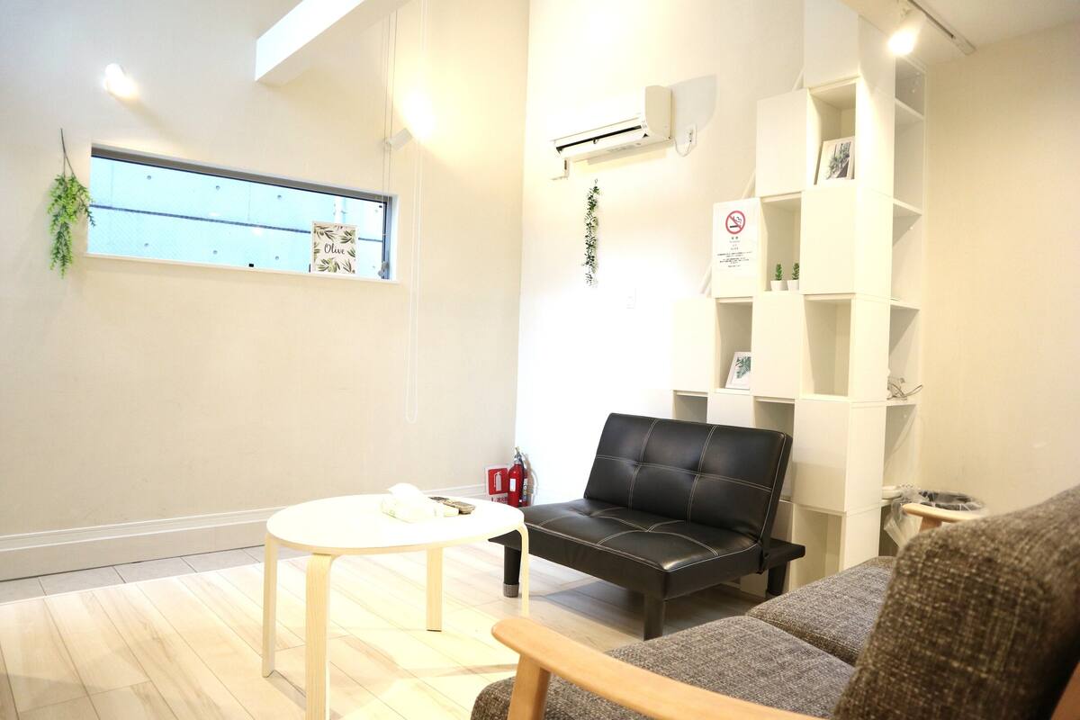 Una sala de estar moderna con un sofá de cuero negro, una mesa de centro blanca y un sillón gris. Se aprecia una estantería blanca decorada con objetos y una pequeña planta en maceta.