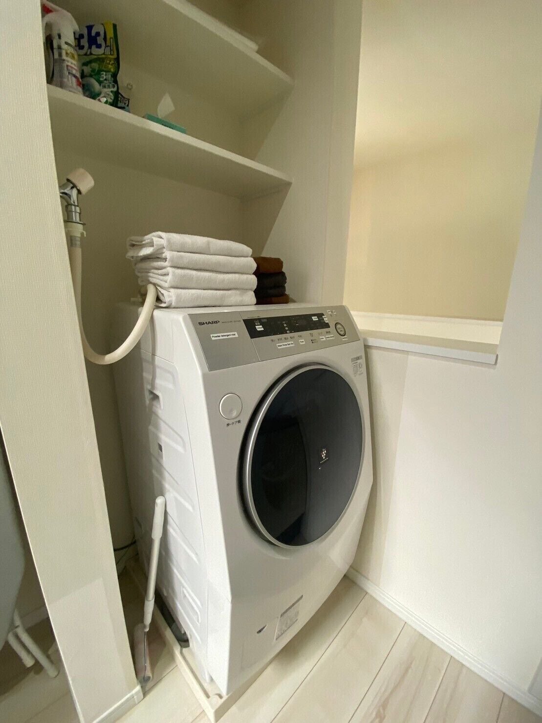 Lavadora blanca de carga frontal, compacta, en un pequeño cuarto de lavado con un estante superior que contiene toallas dobladas y productos de limpieza.