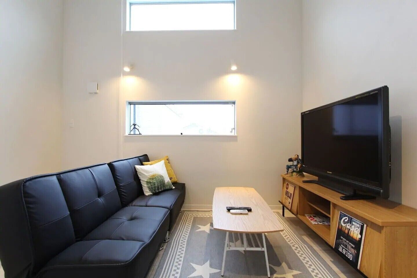 A compact living space with a black leather sofa, a small table, a television on a wooden stand, and a window providing natural light.