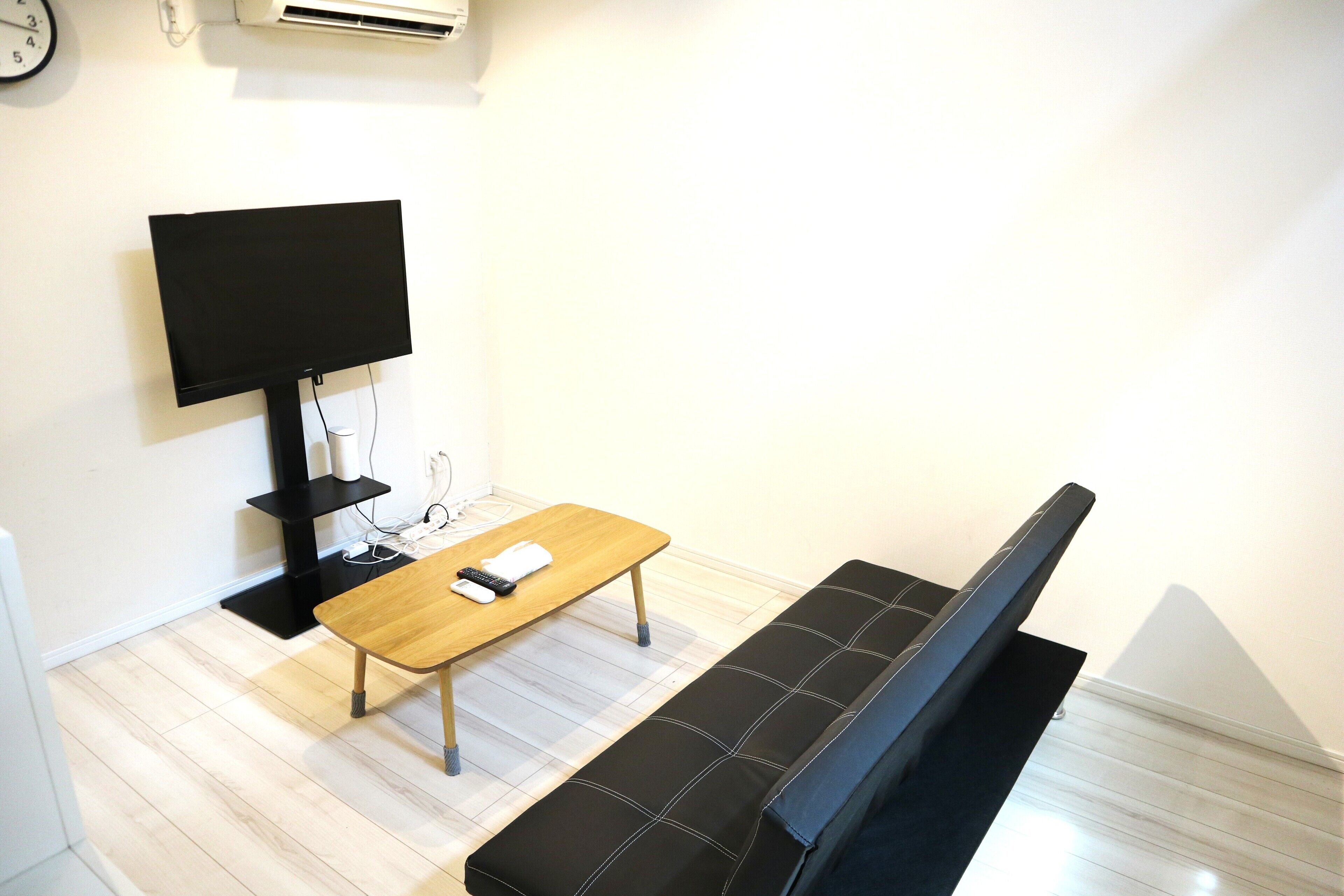 A room with a wooden table, a black sofa, a flat-screen TV mounted on the wall, and a clock on the wall.