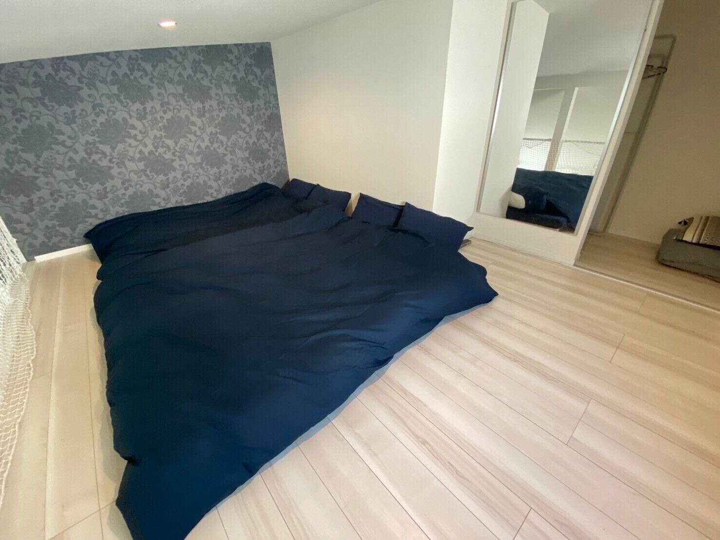 Quarto com cama grande, piso de madeira, parede cinza com padrão e um espelho.