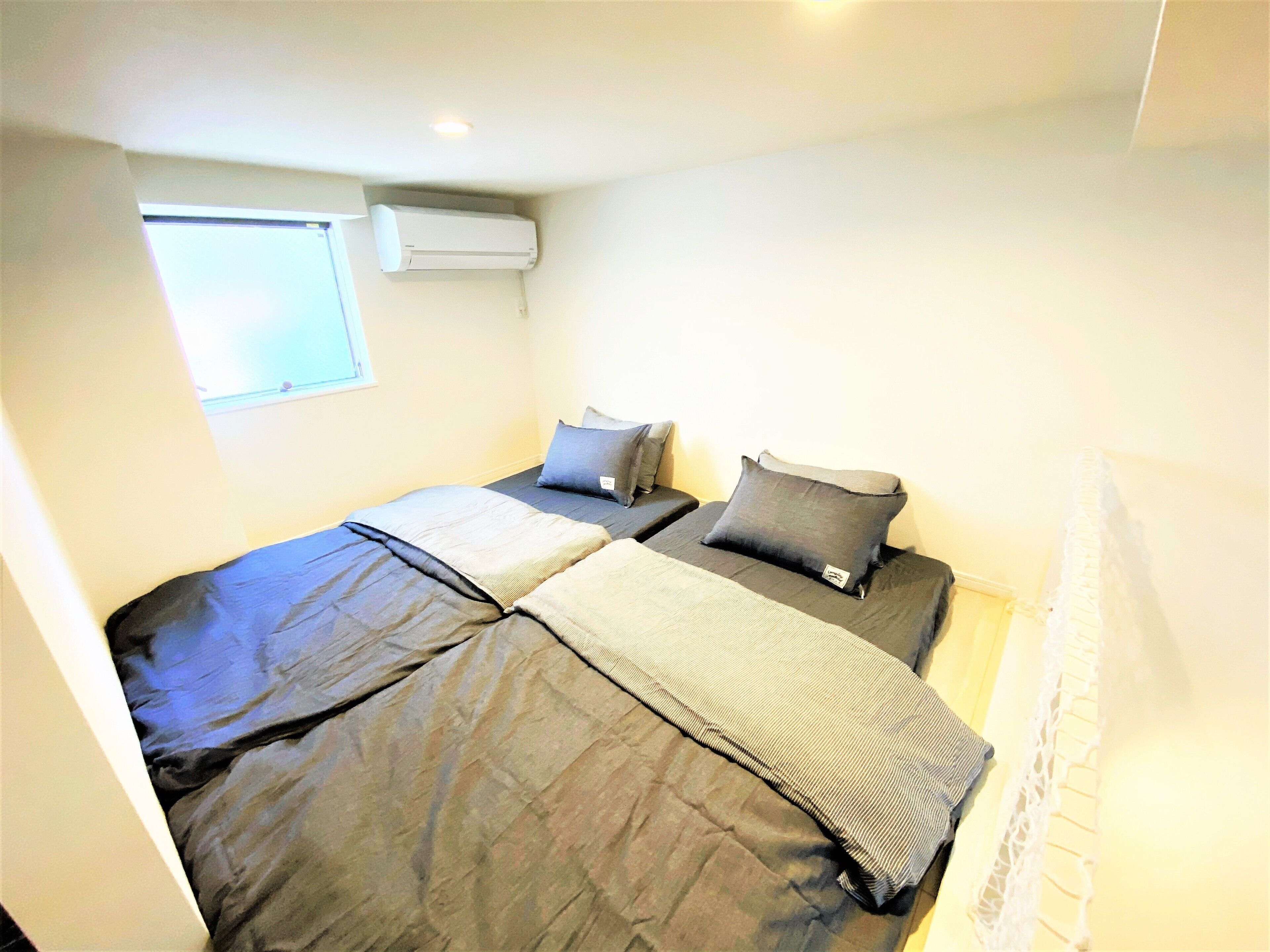A small, compact room with two beds, a window, and an air conditioning unit.