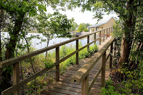 A wooden bridge leading to a building with a sloped roof, surrounded by trees and a body of water. - Villa Oesterdam Sleeps 8 Persons (Tholen)