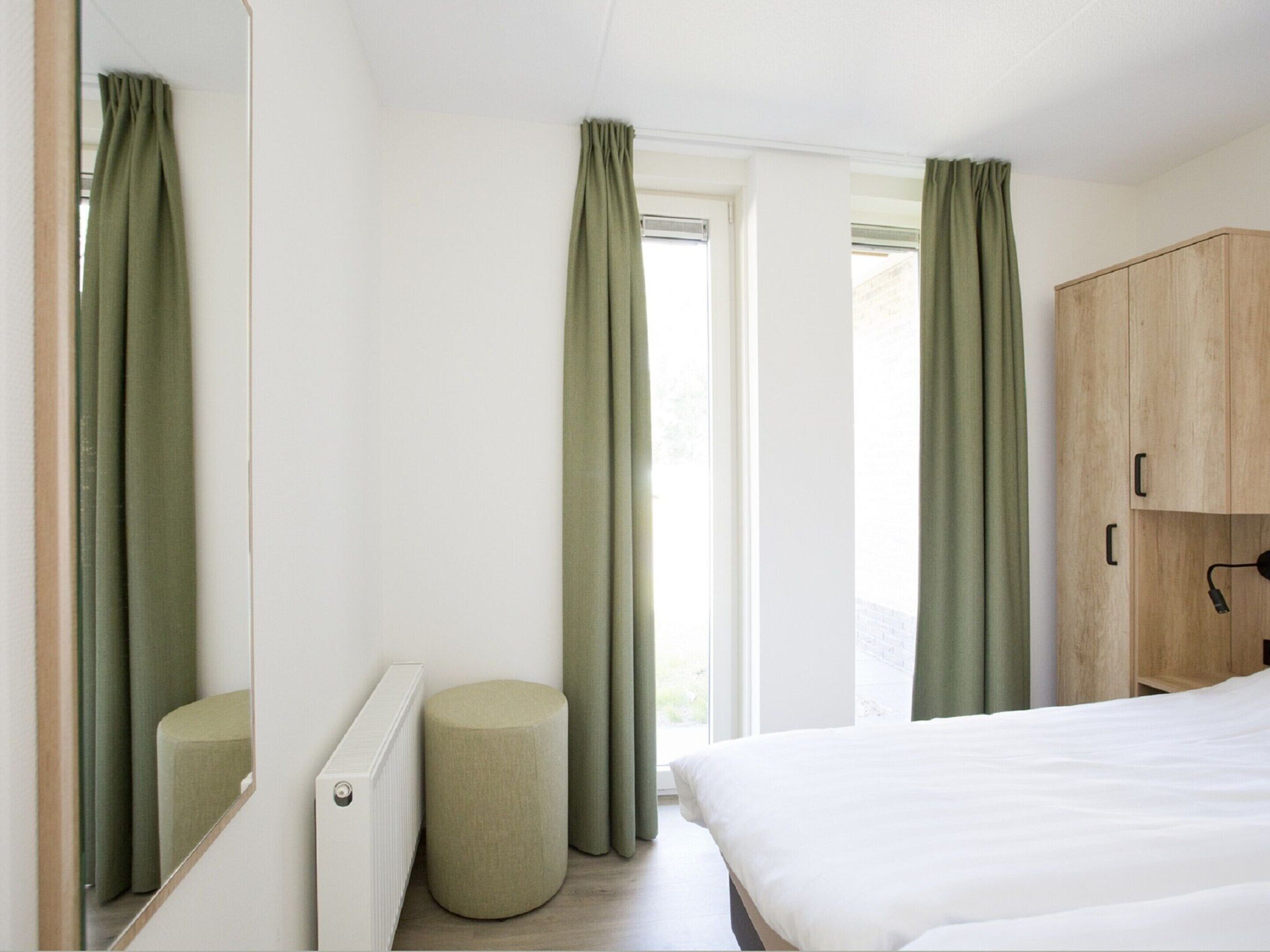 Een slaapkamer met een bed, een houten kledingkast, een rond poef en een raam met gordijnen.