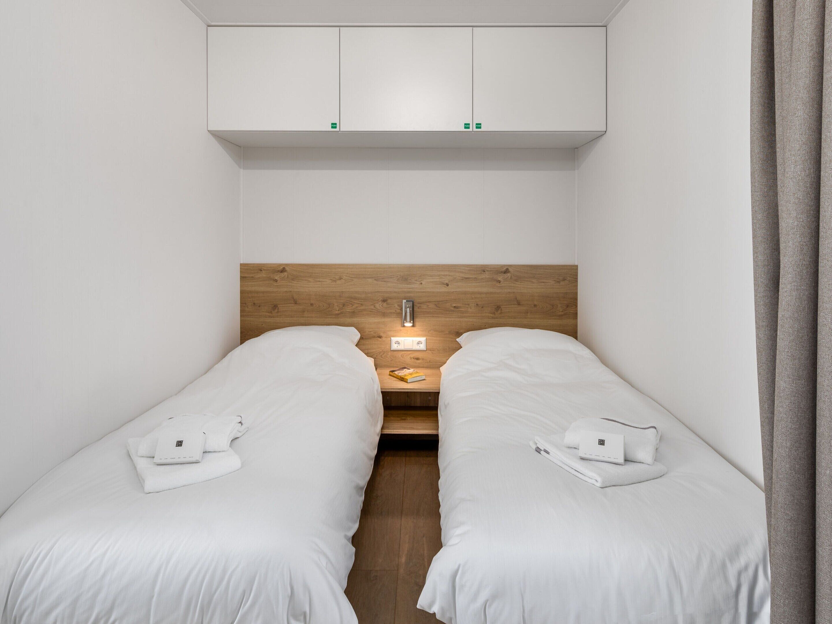A small, modern hotel room with two single beds, a wooden headboard, a nightstand with a lamp, and a white wardrobe above the beds.
